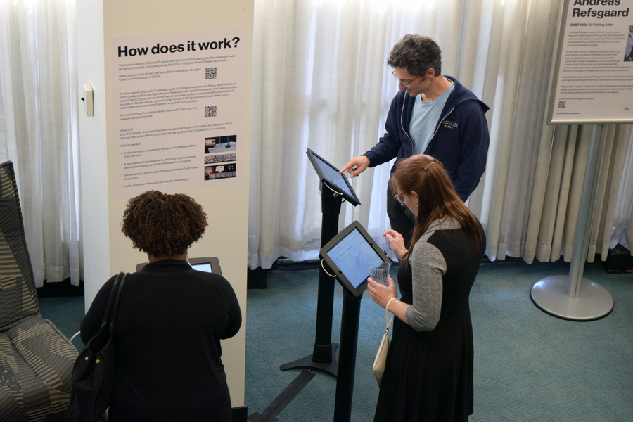 Three people stand in front of a poster labeled “How does it work?” while tapping on pedestal touchscreens.