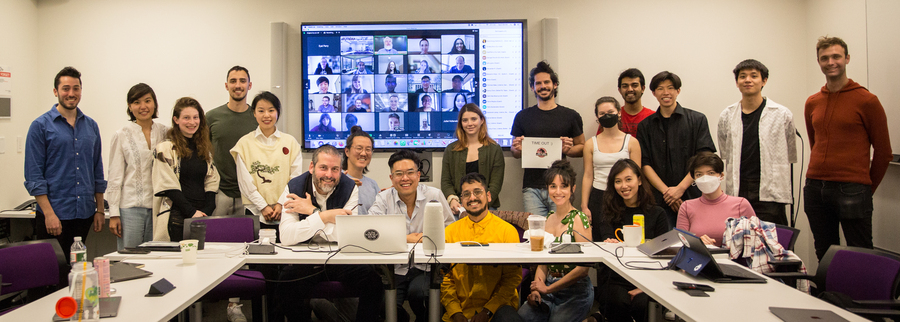 19 people stand and sit around a table posing for a photo. Behind them is a screen with a five by five grid of people on a Zoom call