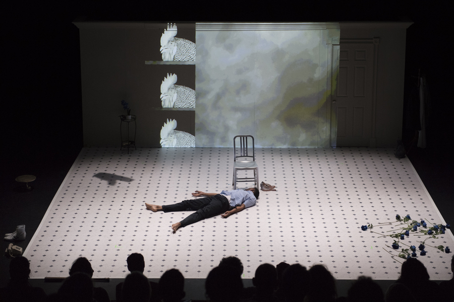 A man lies on his back on stage, next to a chair, with his head turned away from the audience