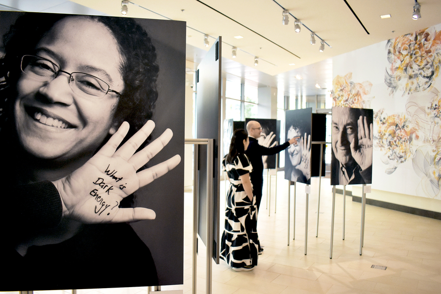A close-up of a grayscale portrait of a woman, with her hand raised up. On her hand is written “What is dark energy?” Beyond her portrait, two people view other images.
