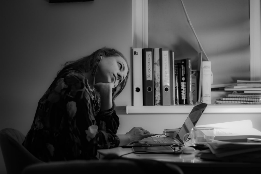 Photo of Tetiana Herasymova sitting at a table with a laptop open in front of her. Her face is forlorn and she's resting her head in her hand. Textbooks are on the shelf behind her.