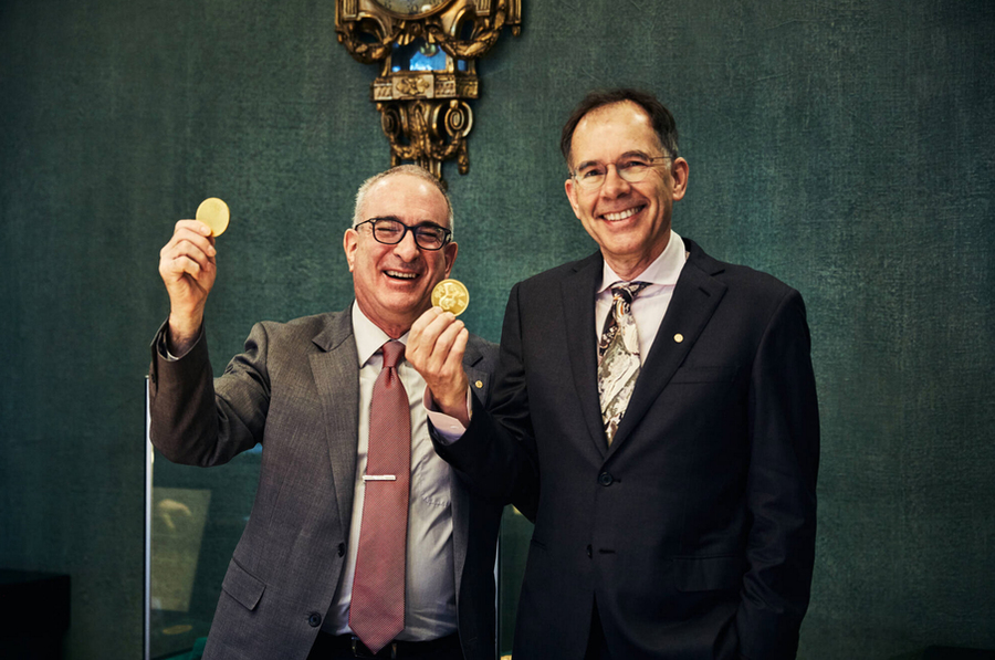 Joshua Agrist and Guido Imbens hold gold Nobel Prize medals indoors against a gray background.