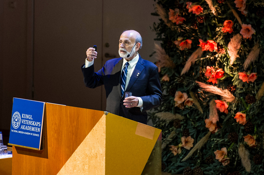 Ben Bernanke gives a speech on stage while holding a small device.