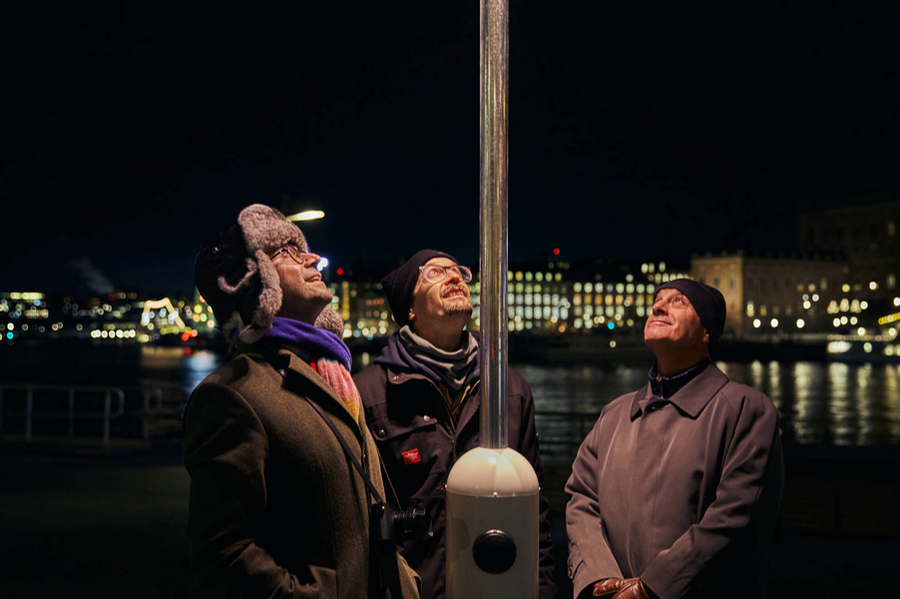 Ardem Patapoutian, David Julius, and Alexander Wolfing look up at a unique street lamp at night with water and city buildings of Stockholm, Sweden, behind them.