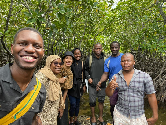 Seven individuals stand and pose for a group photo in a lush green wooded area. 
