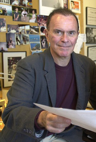 Photo of Richard Wurtman with a sheet of paper in his hand and a bulletin board filled with photos and other memoribilia behind him
