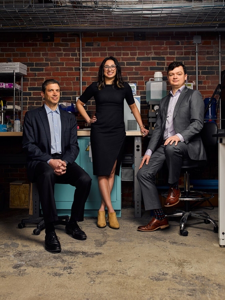 Three people pose in front of a lab bench and a brick wall. Two are seated and one is standing.
