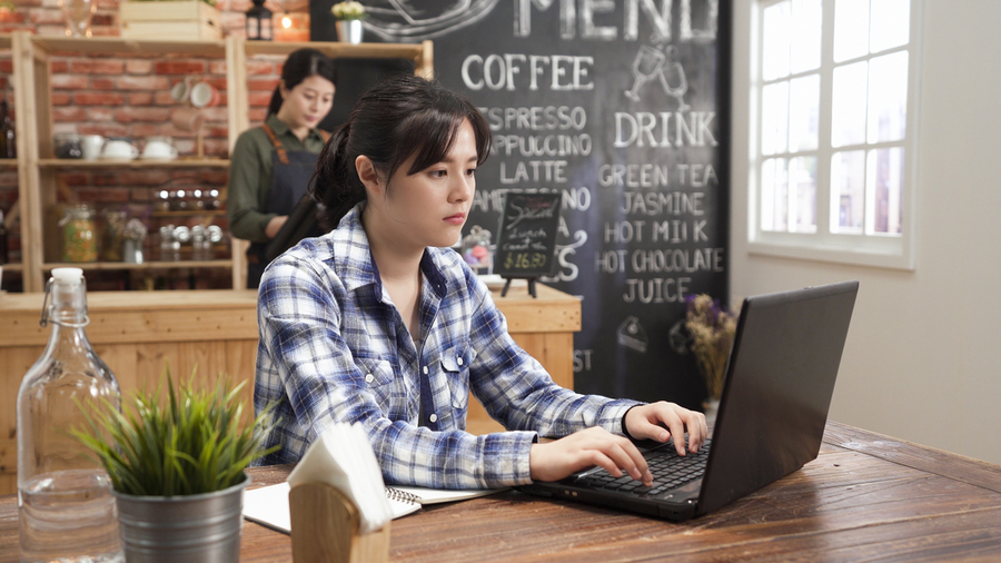 A young woman sits at a cafe table working on her laptop. In the background there is a woman at the counter wearing an apron and a large chalkboard listing the cafe's menu.