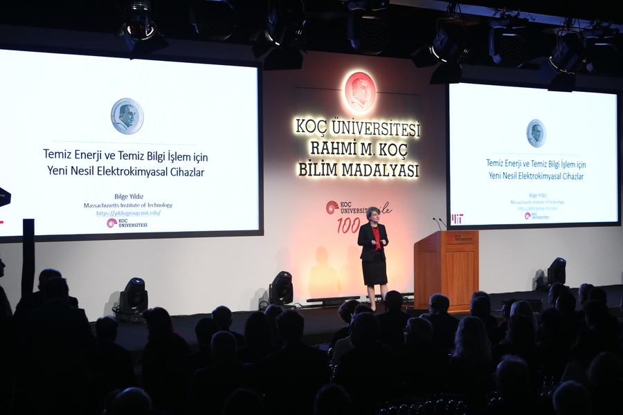 Bilge Yildiz speaks onstage at Koc University in Turkey. There are large screens displaying Turkish text on either side of her.