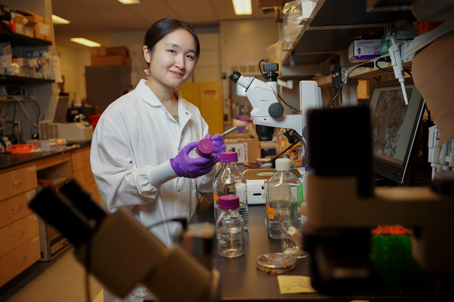 Catherine Ji, in the lab with purple gloves and surrounded by microscopes and lab equipment, labels a glass bottle.