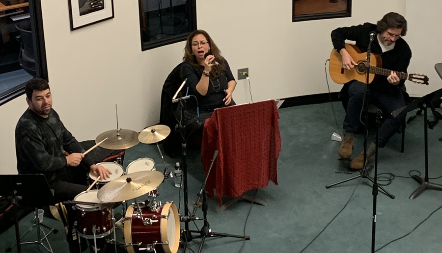 In a white room, Rafael Barata plays drums, Anna Borges sings holding a mic, and Bill Ward plays guitar
