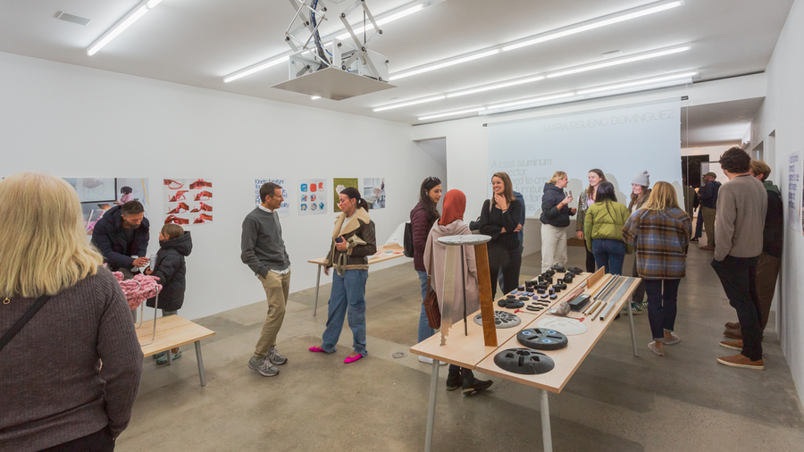 About 15 people mill about in a small gallery space, talking and looking at exhibited items on tables and mounted on walls.
