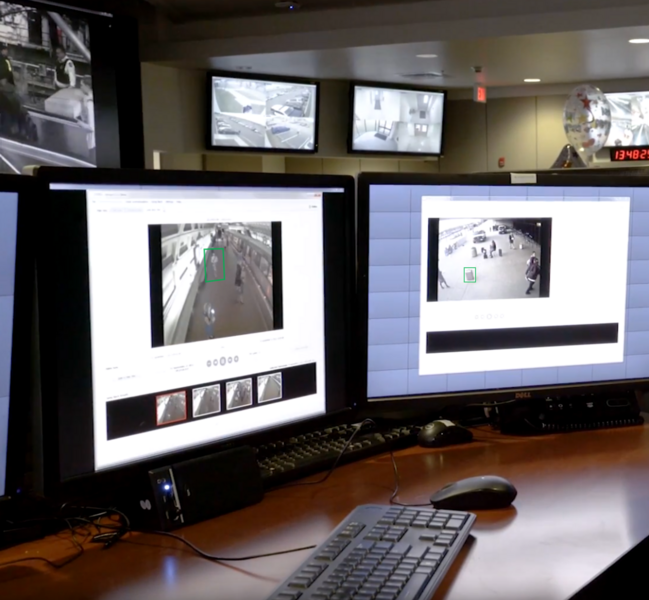 In the foreground, two desktop monitors display surveillance videos of a subway and a street scene. Two more monitors are mounted near the ceiling in the background