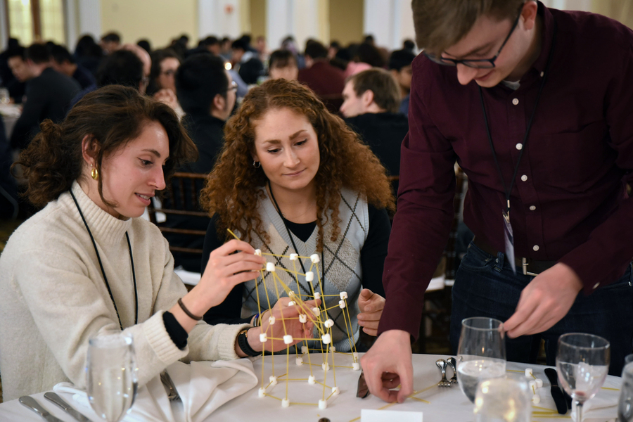 Three MIT graduate students building a lattice-like structure at a dinner table using spaghetti and mini marshmallows
