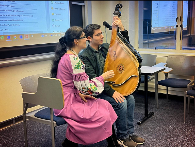 A woman in traditional Ukrainian dress instructing a student holding a large stringed instrument