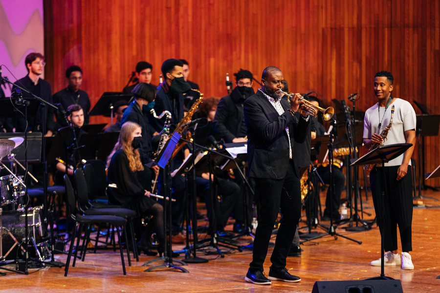 Sean Jones solos on trumpet next to Braxton Cook, who holds a saxophone, while members of a jazz ensemble watch on behind them.