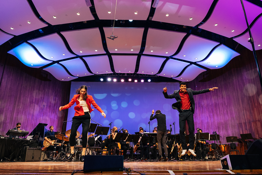 Two dancers jump in midair on stage at MIT's Kresge Auditorium. An orchestra plays behind them.