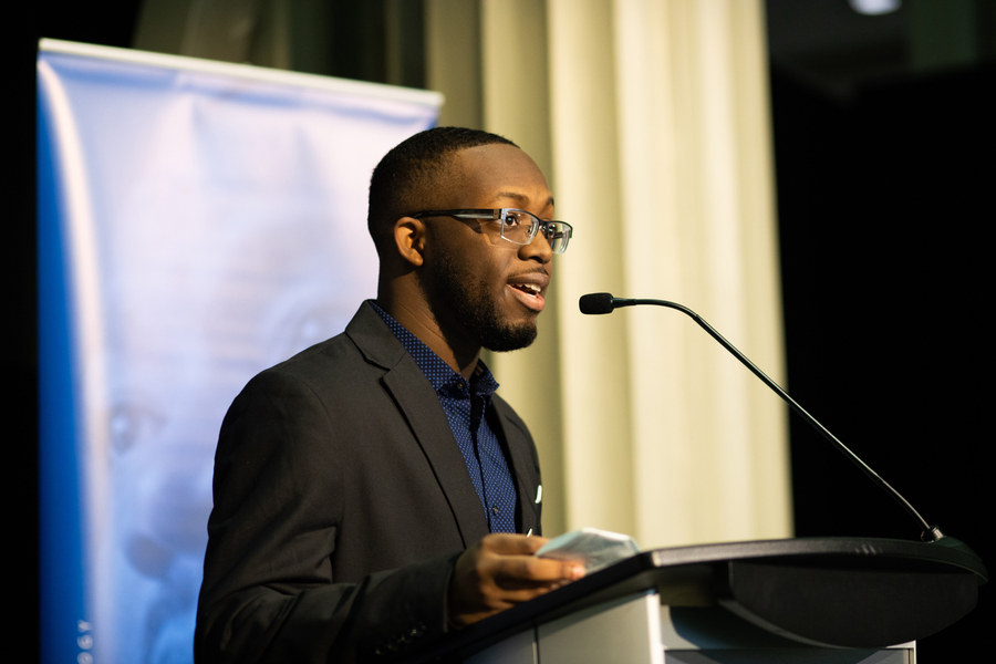 Myles Noel speaks at the podium with an out-of-focus blue banner in background.