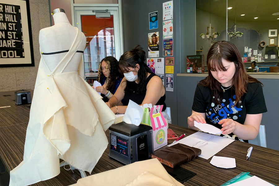 Three students work at a table, with small mannequin draped in plain white fabric placed in front of them.