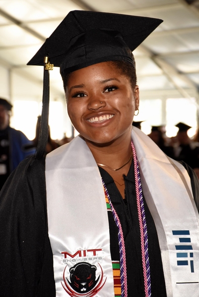 Elissa Gibson dressed in her MIT regalia during commencement. The photo is taken from the middle of her torso up. She is wearing a black graduation gown, black graduation cap with black tassel, white sash with “MIT Engineers” and a beaver embroidered on the left, and MIT logo in blue on the right. She wears three cords, one red and white, one purple and white, and one woven with black, green, red, white, and yellow alternating. She is standing in a large white outdoor tent, other graduates can be seen blurr