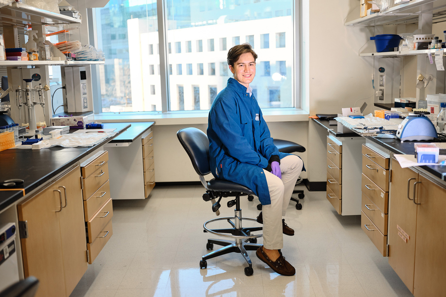 Julian Zulueta, wearing a blue lab coat and purple gloves, sits on a chair in a lab with a large window in background.