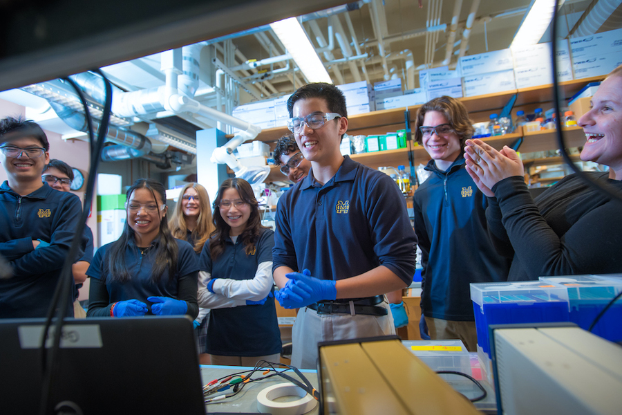 Eight high school students standing in lab, smiling at a monitor.