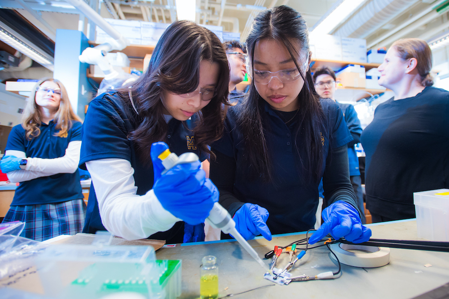 Brianna Tong and Angelina Ang lean over a workbench, adding an electroactive solution to their electrode with a large pipette