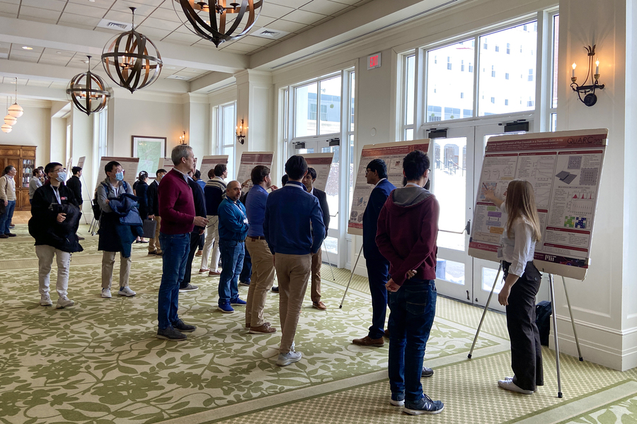 Poster sessions at QuARC 2023: A row of MIT graduate students and postdocs stand in front of their posters while visitors walk by and stop to listen.