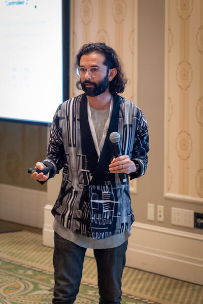 Erik Lucero standing and speaking before an audience. He has a microphone in one hand and a slideshow clicker in the other.
