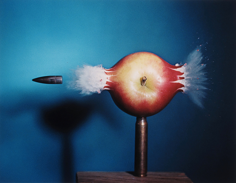 A bullet shoots through an apple, leaving clouds of ejecta on either side.