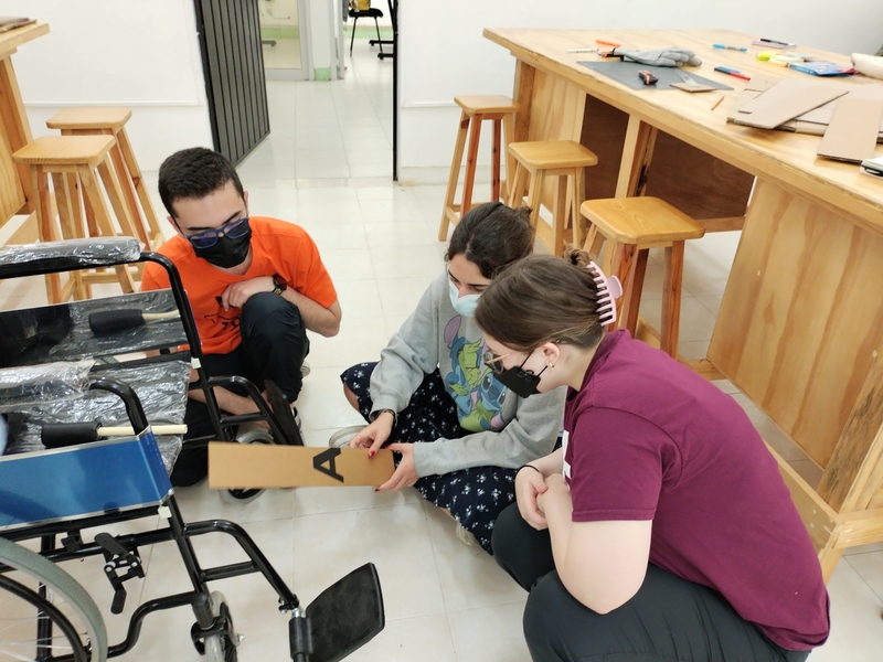 Three MIT students sit on the floor, examining an unoccupied wheelchair