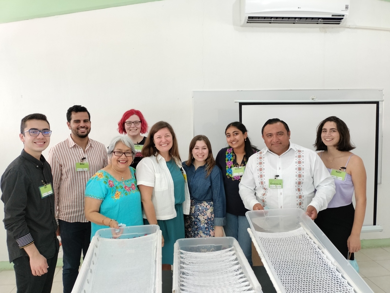 Nine people pose in a line in a white room. Three white prototype bathtubs stand before them