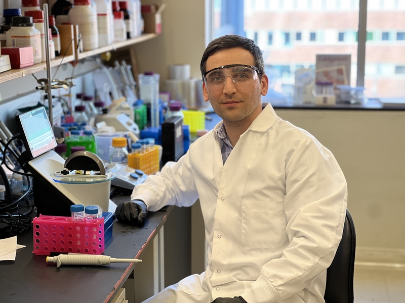 Amir Erfani in a lab coat and protective gear, sits at a lab bench