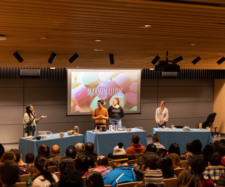 Four presenters stand behind three tables with cooking supplies in an auditorium