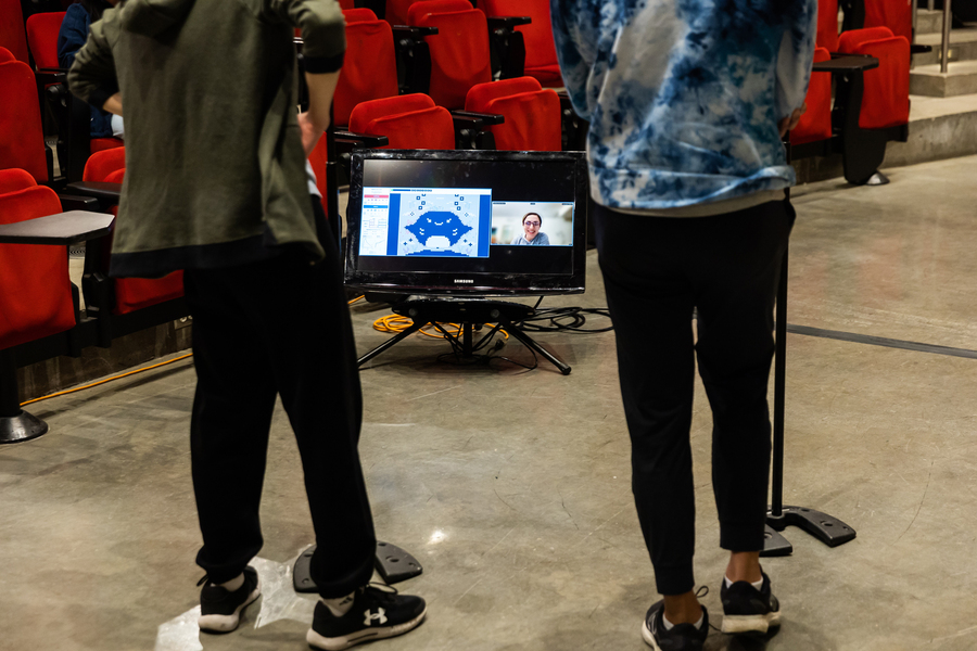 A monitor displaying split screen view shows a virtual battlefield and an individual as it faces two individuals.