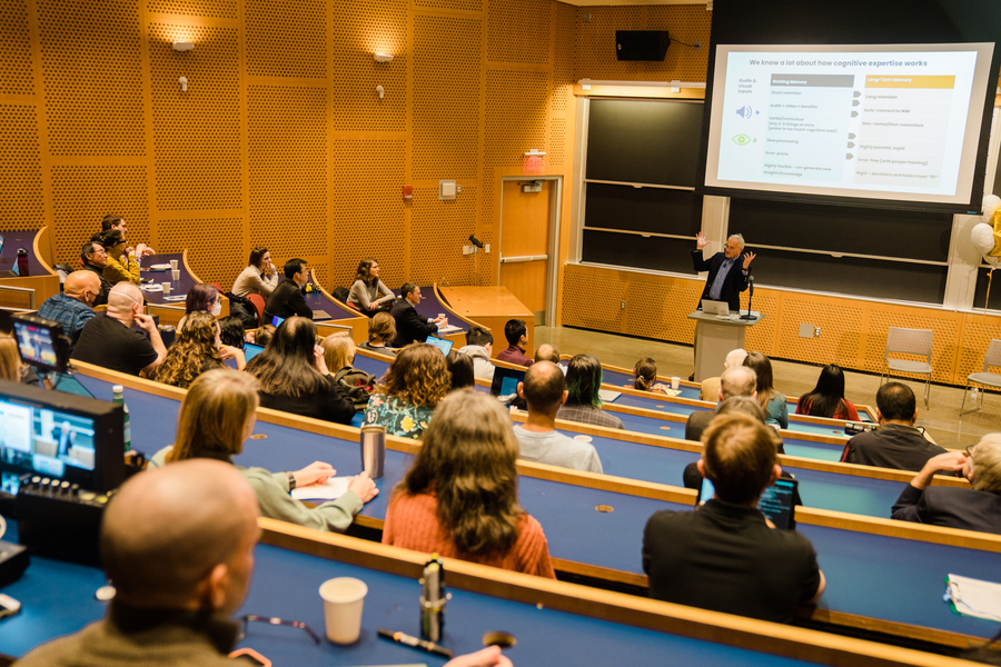 Photo taken from the back of a college lecture hall showing several dozen adults listen to a speaker who is standing between a podium and a screen displaying a slideshow.