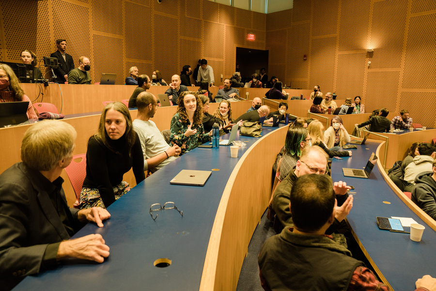 Photo of several dozen adults seated in a college lecture hall, engaged in conversations with each other. Audience members look to be excited, smiling, and listening intently.