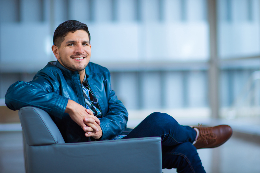 Vila sits on a blue chair with legs crossed, and large windows in background.