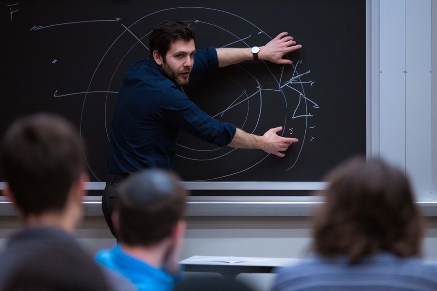Jacob Andreas, at a blackboard, points to a diagram in chalk. The backs of student's heads are in the foreground