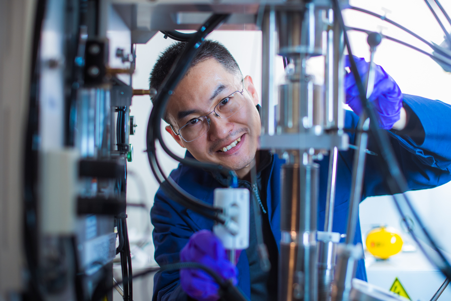 Mingda Li, wearing nitrile gloves, smiles behind equipment in lab.