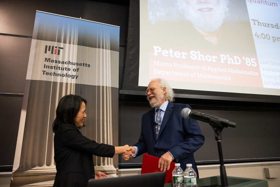 Lily Tsai shakes hands with Peter Shor on the presentation stage. Shor is holding a red folder, and both are smiling.