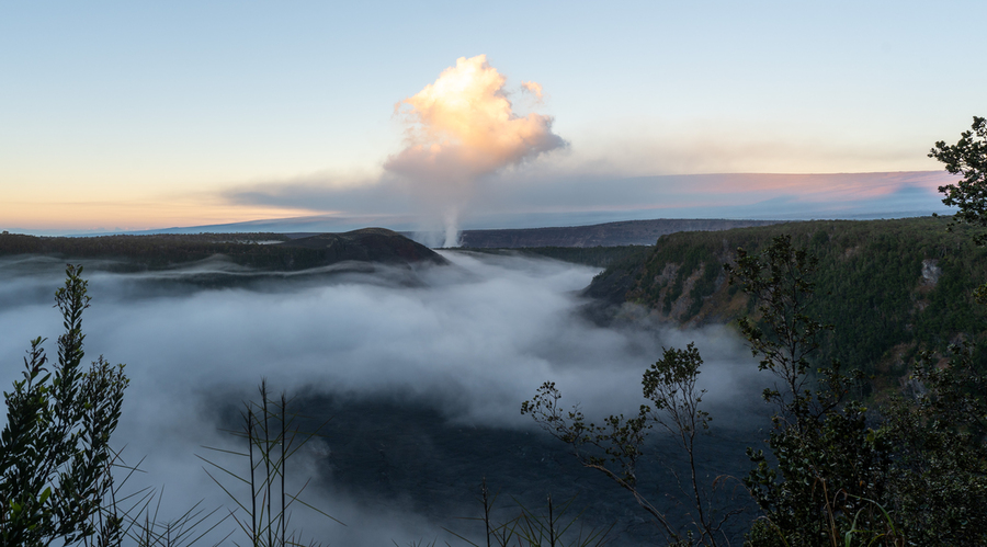 Volcanic plume against a blue sky with mist accumulation building around a mountain