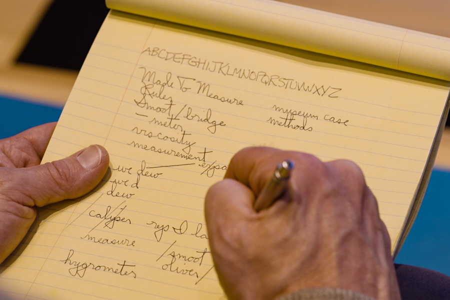 Closeup of Duncan’s yellow notepad and his hands as he writes in cursive. The alphabet is on top of the page, and other words appear: “Made To Measure; Museum Case. Methods.” 