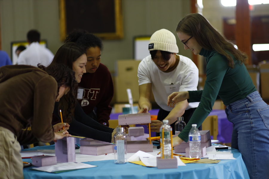 Students laugh and stand around a table while building a model skyscraper with foam and pencils.