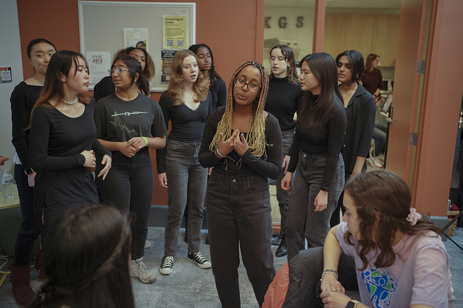 Group of 10 women, all wearing black, singing in MIT's Cheney Room while others look on