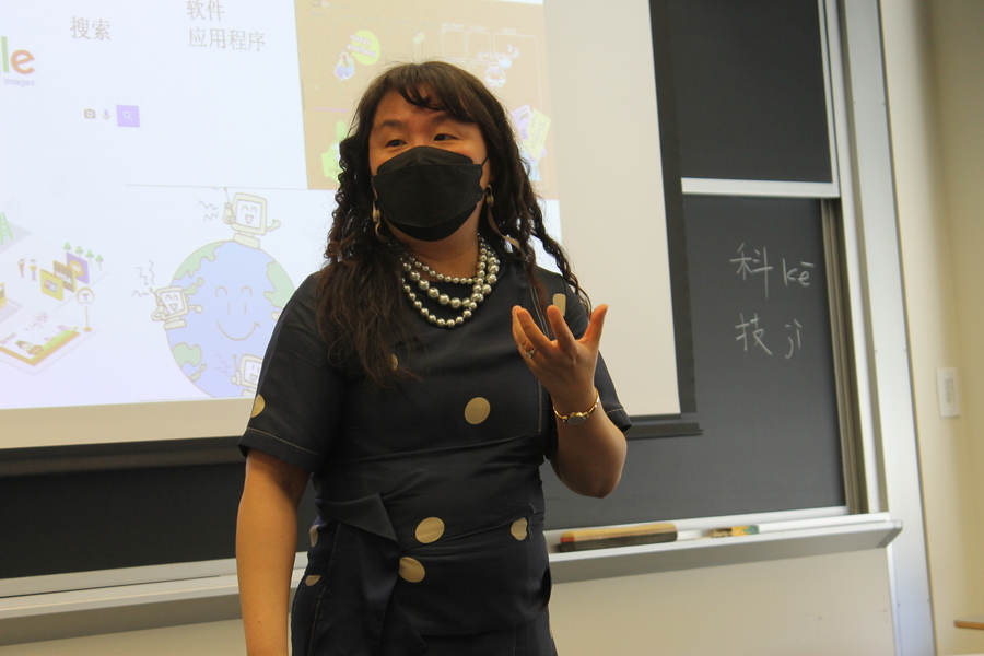 A woman in a polka-dot dress gestures while standing in front of a projection screen. 