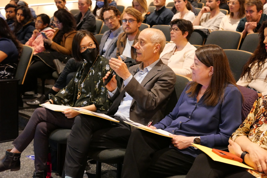 Seated, with papers on his lap, Bruce Birren gestures, holding a microphone, as he speaks. Other judges and attendees are seated around him.