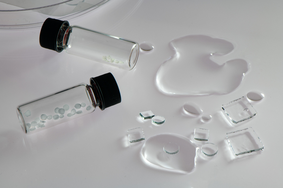 On a white table, 2 small glass vials contain pebble-like translucent chips. Some puddles of water are on the table, as well as circular and square chips of a hydrogel substance.