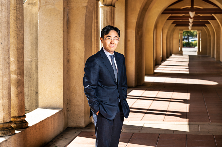 In Song Kim stands and smiles while outdoors on a sunny day at Stanford University. In the background is a long walkway with arches.