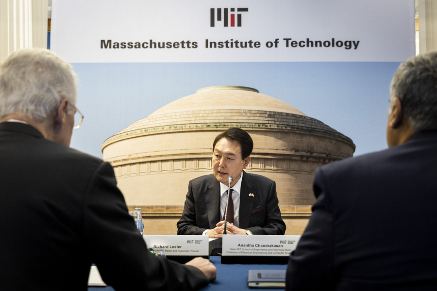 President Yoon Suk Yeol sits at a table in front of a microphone, speaking. Two people are facing him, with their backs to the camera, and behind him is a screen showing a photo of MIT’s Great Dome, with the MIT logo and the words “Massachusetts Institute of Technology.”